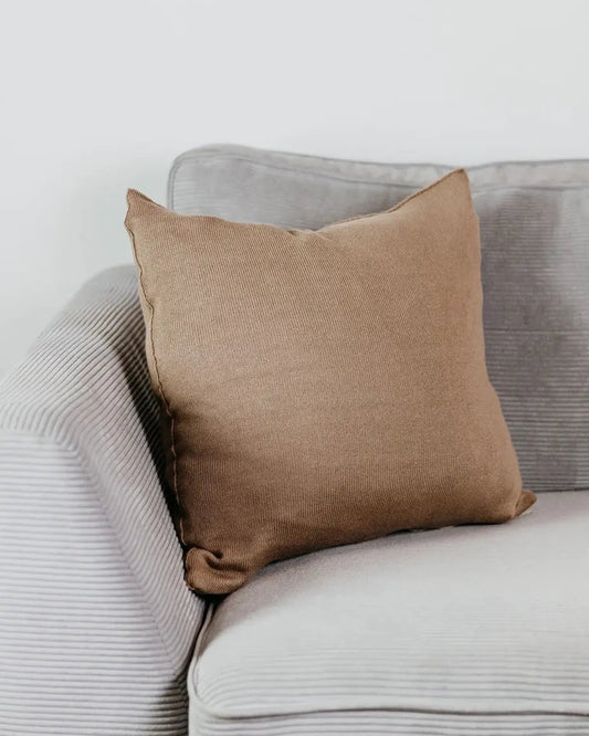Beiges Strick-Dekokissen auf einem hellgrauen Sofa vor einer weißen Wand.