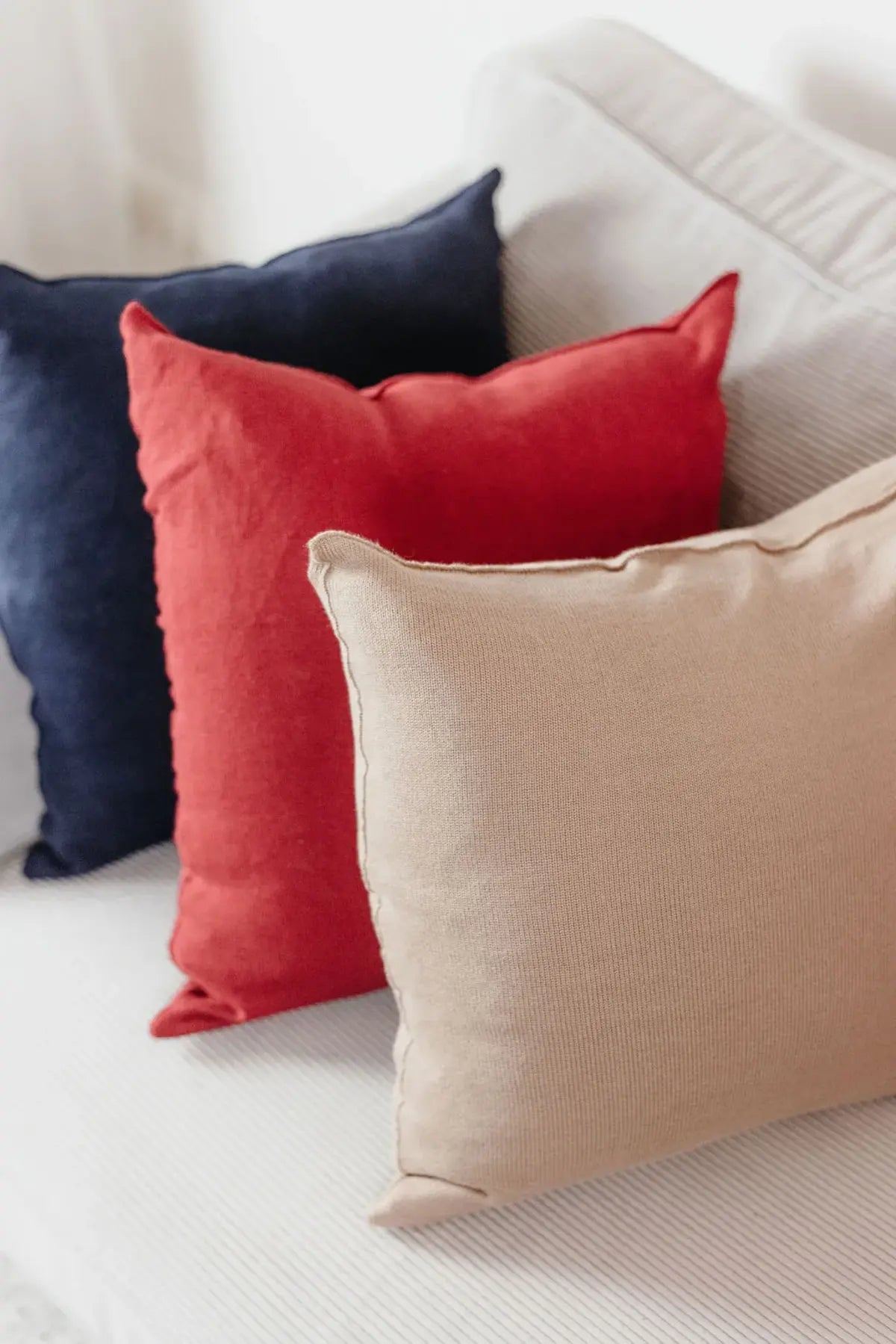 Drei Strick-Dekokissen in Navyblau, Weinrot und Cremebeige auf einem hellen Sofa arrangiert.