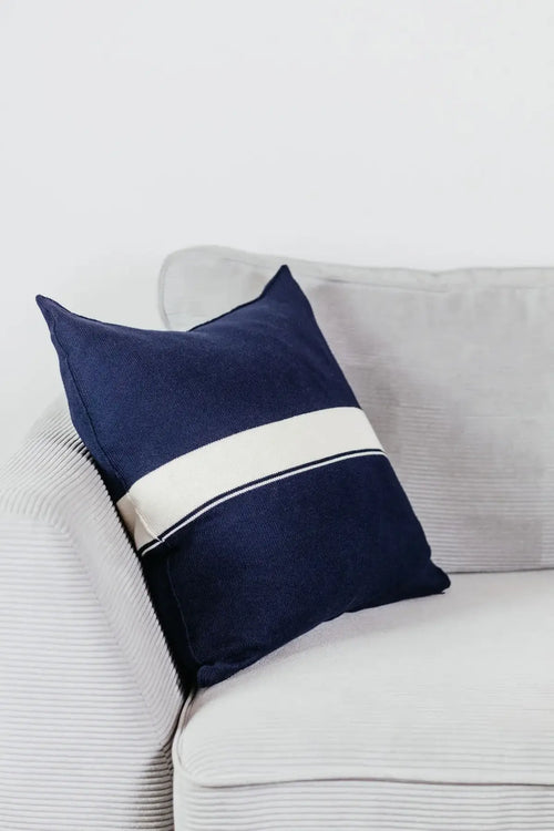 Navyblaues Strick-Dekokissen mit cremefarbenem Streifen auf einem hellgrauen Sofa vor einer weißen Wand.