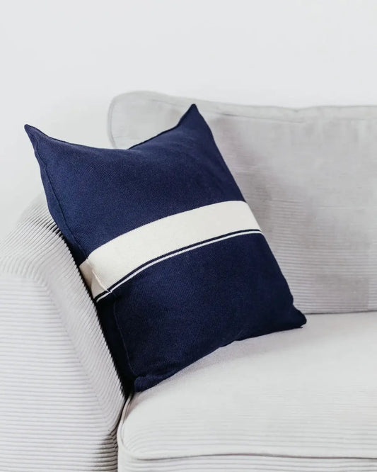 Navyblaues Strick-Dekokissen mit cremefarbenem Streifen auf einem hellgrauen Sofa vor einer weißen Wand.
