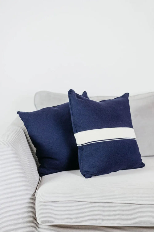 Zwei Strick-Dekokissen in Navyblau einfarbig und Navyblau mit cremefarbenem Streifen auf hellgrauem Sofa im Wohnbeispiel.