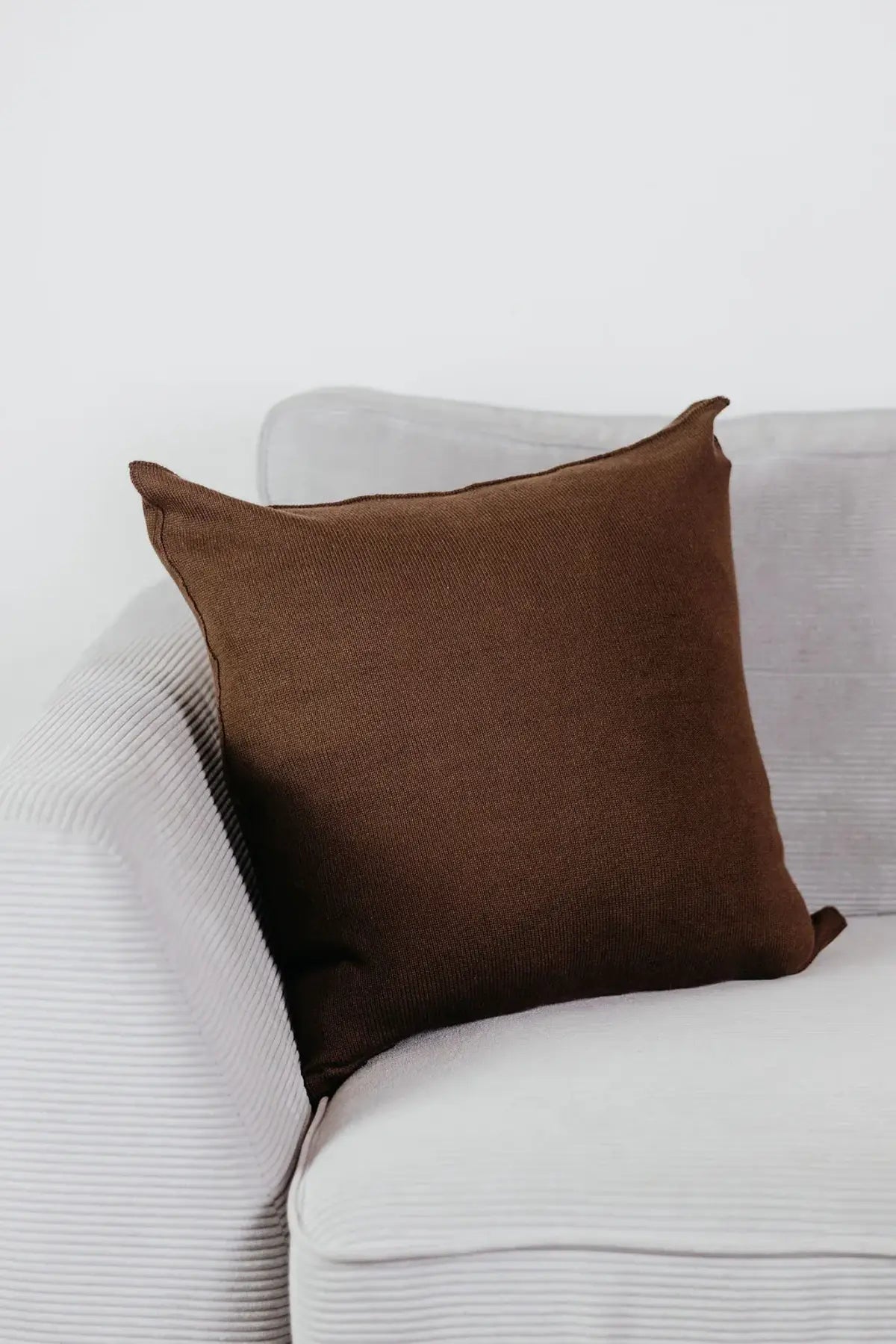 Braunes Strick-Dekokissen auf einem hellgrauen Sofa vor einer weißen Wand.