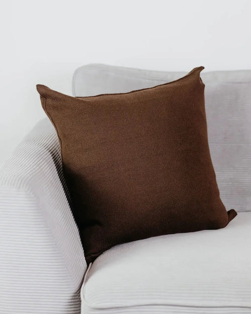 Braunes Strick-Dekokissen auf einem hellgrauen Sofa vor einer weißen Wand.
