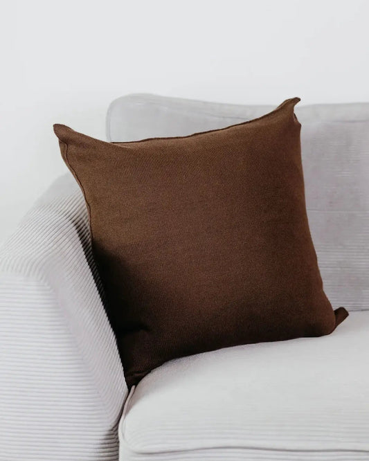 Braunes Strick-Dekokissen auf einem hellgrauen Sofa vor einer weißen Wand.