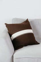 Braunes Strick-Dekokissen mit cremefarbenem Streifen auf einem hellgrauen Sofa vor einer weißen Wand.