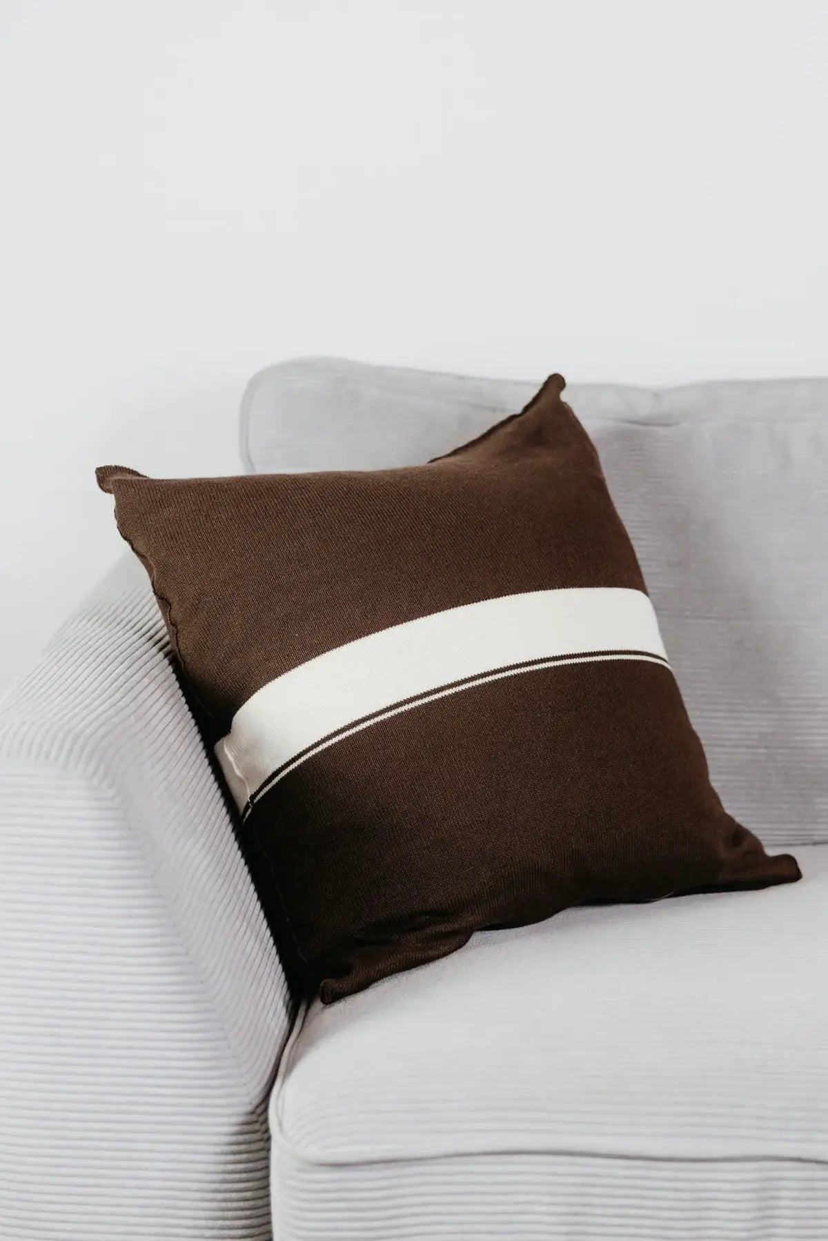Braunes Strick-Dekokissen mit cremefarbenem Streifen auf einem hellgrauen Sofa vor einer weißen Wand.