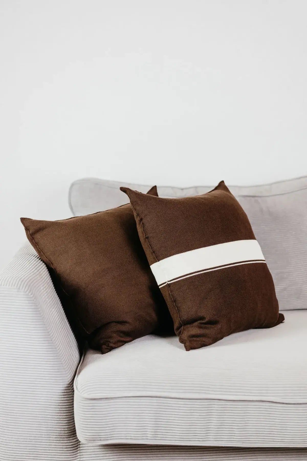 Zwei Strick-Dekokissen in Braun einfarbig und Braun mit cremefarbenem Streifen auf hellgrauem Sofa im Wohnbeispiel.