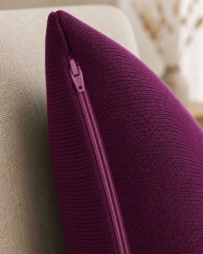 Detailansicht des Reißverschlusses an der Seite eines violetten, gestrickten Dekokissens, das auf einem beigen Sessel liegt.