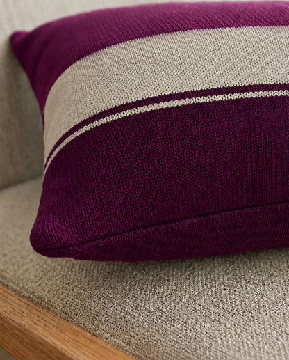 Nahaufnahme eines violetten Dekokissens mit gestricktem hellen Streifen, das auf einem beigefarbenen Polstermöbel mit Holzrahmen liegt.