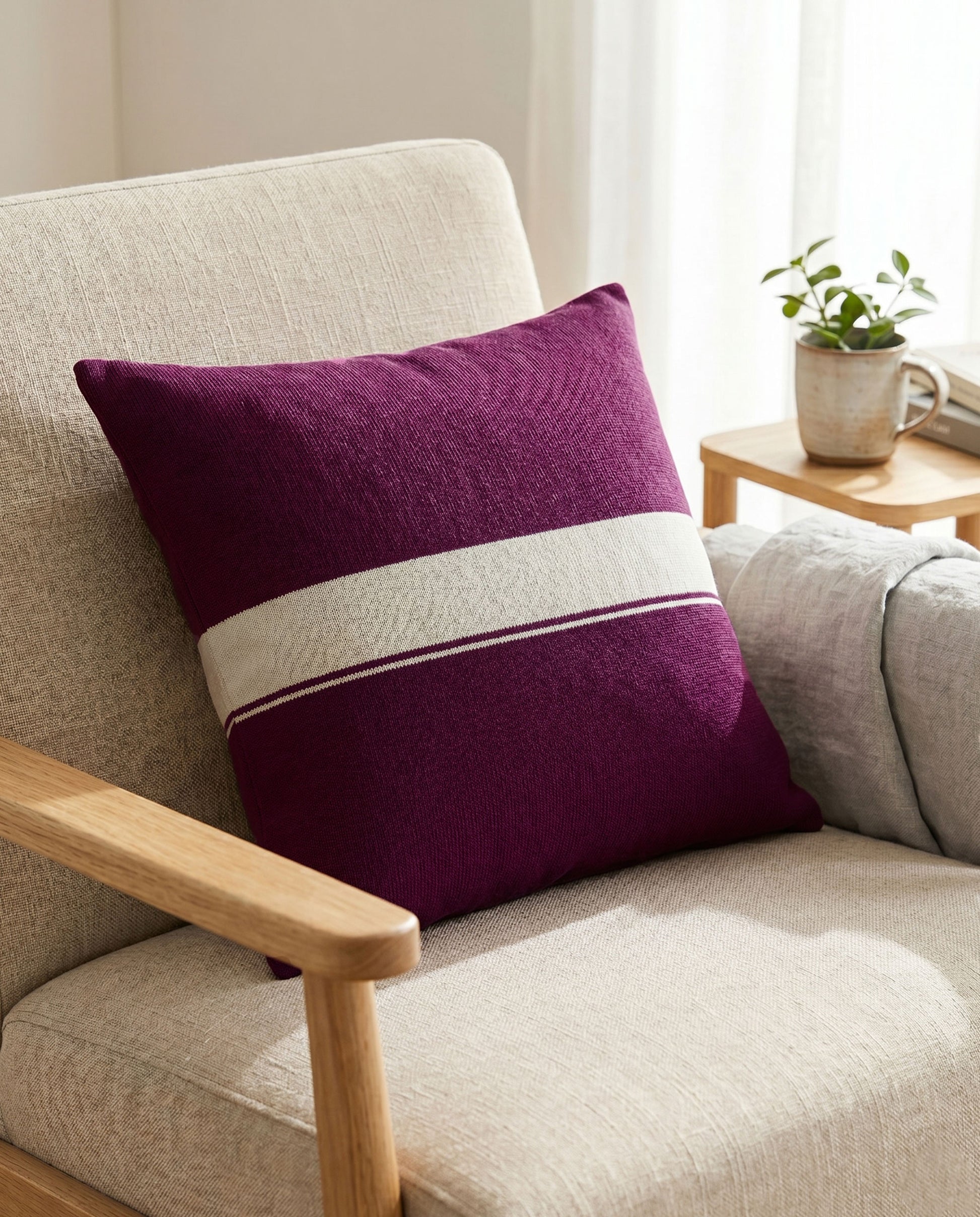 Ein violettes Dekokissen mit dem hellen Streifen in der Mitte, platziert auf einem hellen Polstersessel neben einem Holztisch.