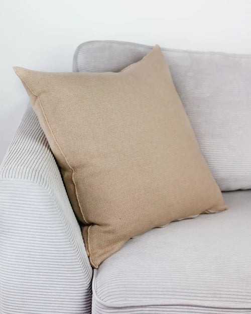 Cremebeiges Strick-Dekokissen auf einem hellgrauen Sofa vor einer weißen Wand.