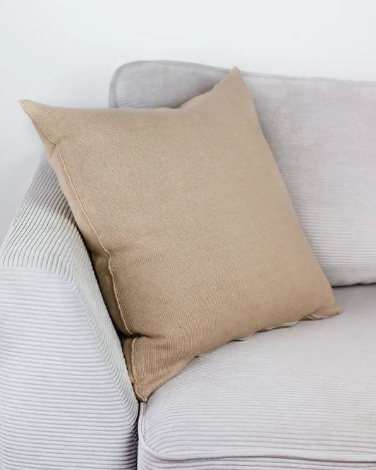 Cremebeiges Strick-Dekokissen auf einem hellgrauen Sofa vor einer weißen Wand.