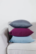 Drei Strick-Dekokissen in Grau, Beerenfarben und Hellblau auf einem Sofa gestapelt.