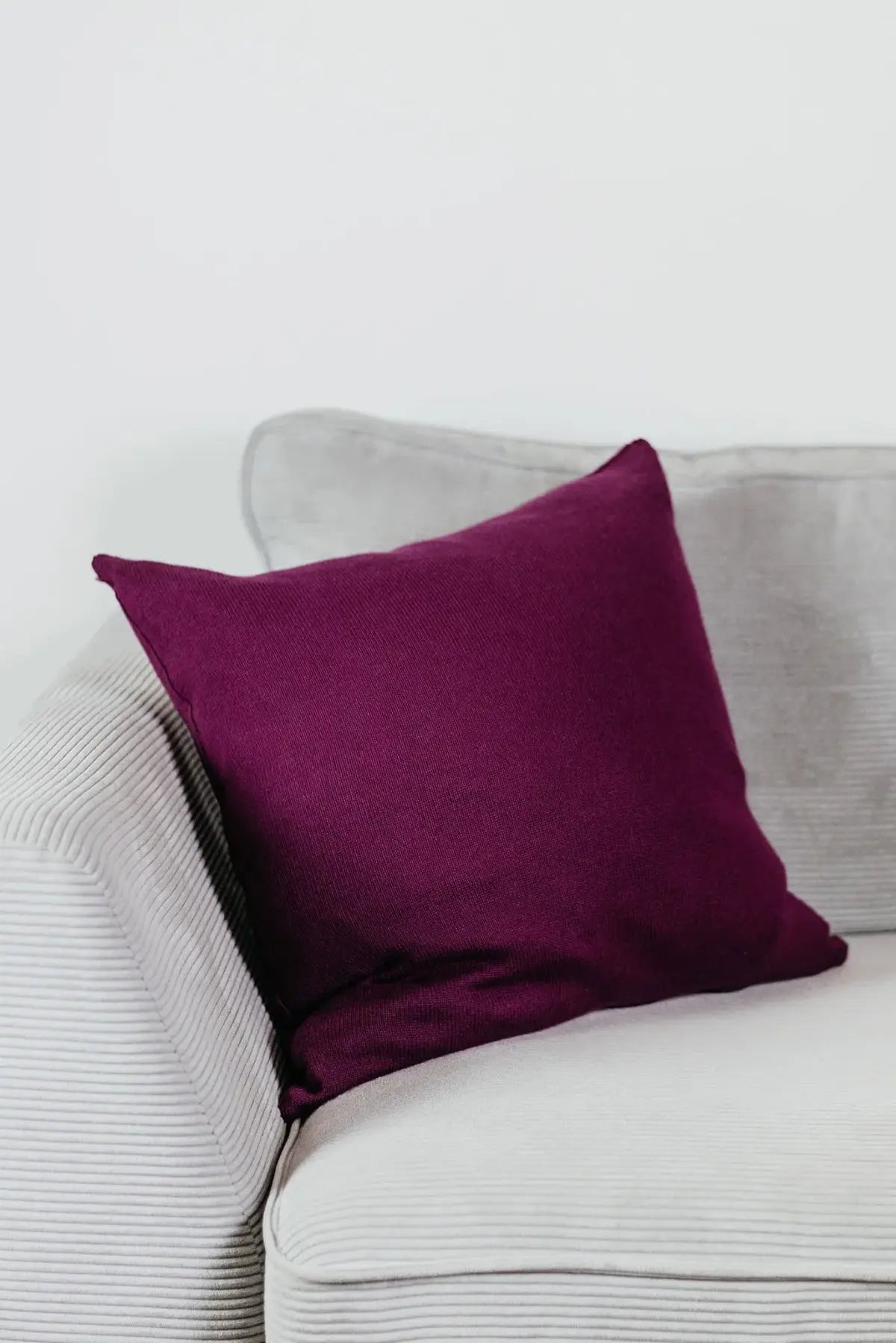 Beerenfarbenes Strick-Dekokissen auf einem hellgrauen Sofa vor einer weißen Wand.