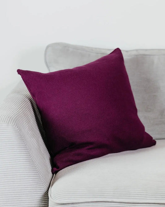 Beerenfarbenes Strick-Dekokissen auf einem hellgrauen Sofa vor einer weißen Wand.