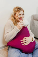 Lächelnde Frau sitzt auf dem Boden und hält ein beerenfarbenes Strick-Dekokissen in der Hand.