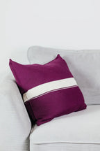 Beerenfarbenes Strick-Dekokissen mit cremefarbenem Streifen auf einem hellgrauen Sofa vor einer weißen Wand.