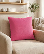 Pinkes Dekokissen auf einem beigen Sessel in einem warm eingerichteten Wohnzimmer mit Holzregal und Zimmerpflanze im Hintergrund.
