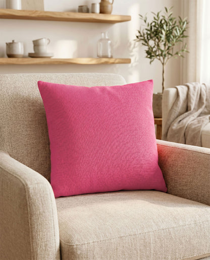 Pinkes Dekokissen auf einem beigen Sessel in einem warm eingerichteten Wohnzimmer mit Holzregal und Zimmerpflanze im Hintergrund.