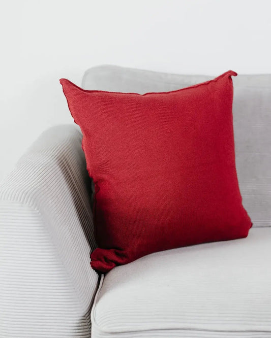 Weinrotes Strick-Dekokissen auf einem hellgrauen Sofa vor einer weißen Wand.