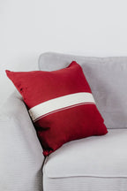 Weinrotes Strick-Dekokissen mit cremefarbenem Streifen auf einem hellgrauen Sofa vor einer weißen Wand.