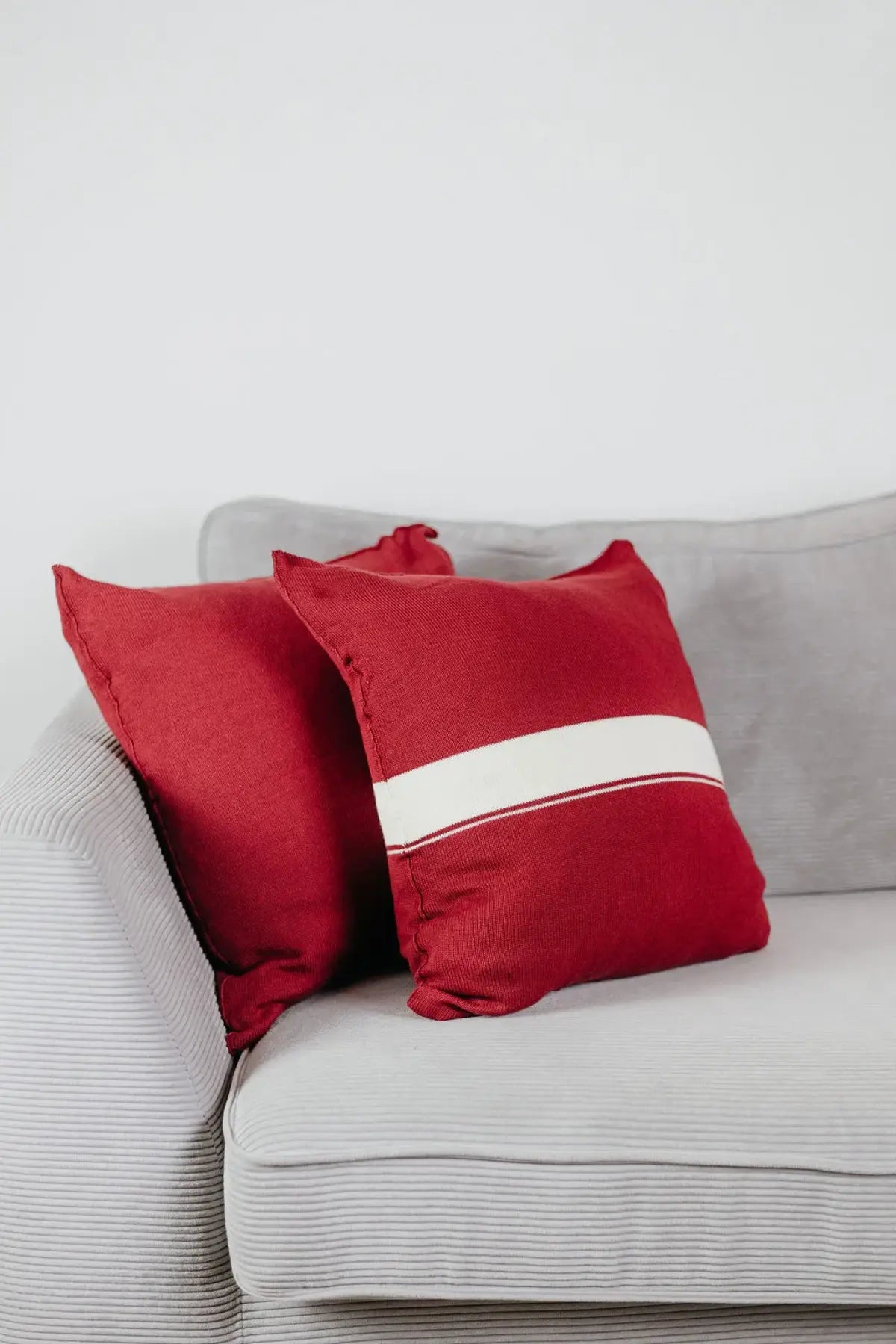 Zwei Strick-Dekokissen in Weinrot einfarbig und Weinrot mit cremefarbenem Streifen auf hellgrauem Sofa im Wohnbeispiel.