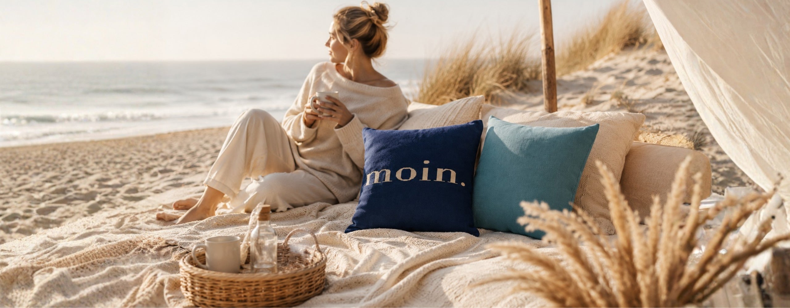 KI-generierte Kulisse: Eine Frau sitzt entspannt an einem sonnigen Strand auf einer Decke mit Kissen, darüber ein dunkelblaues Kissen mit dem Schriftzug „moin.“.