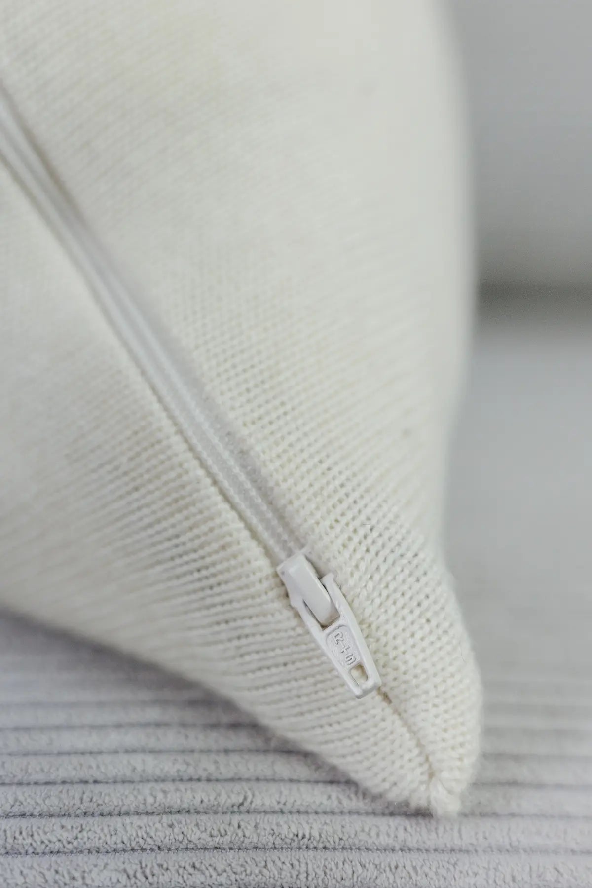 Detailaufnahme eines cremeweißen Strick-Dekokissens mit eingenähtem Reißverschluss auf einem hellen Sofa.