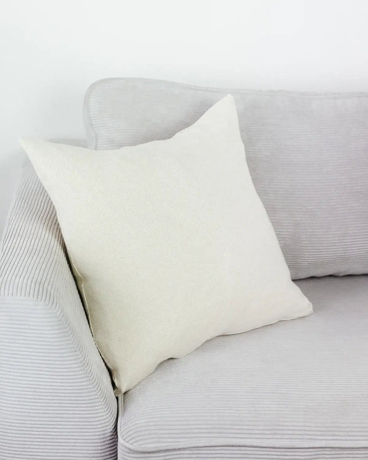 Cremeweißes Strick-Dekokissen auf einem hellgrauen Sofa vor einer weißen Wand.