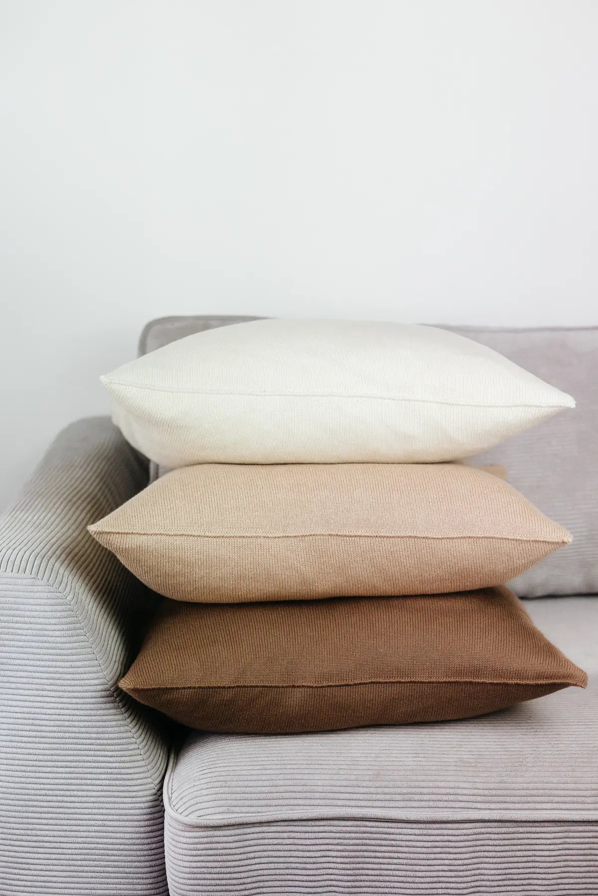 Drei Strick-Dekokissen 50x50 cm in Cremeweiß, Cremebeige und Braun auf einem Sofa gestapelt.