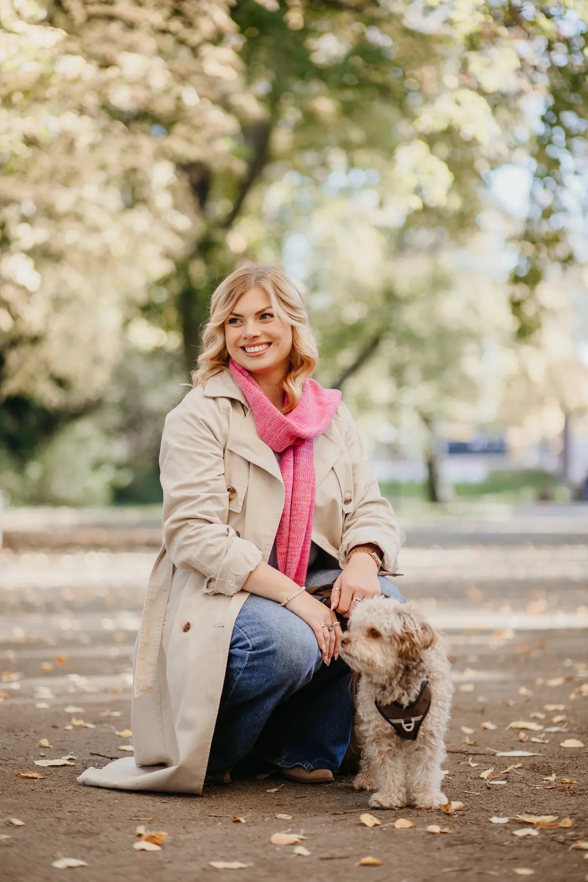 Frau trägt Strickschal in Rosa-Melange im Park und kniet neben einem kleinem Hund.